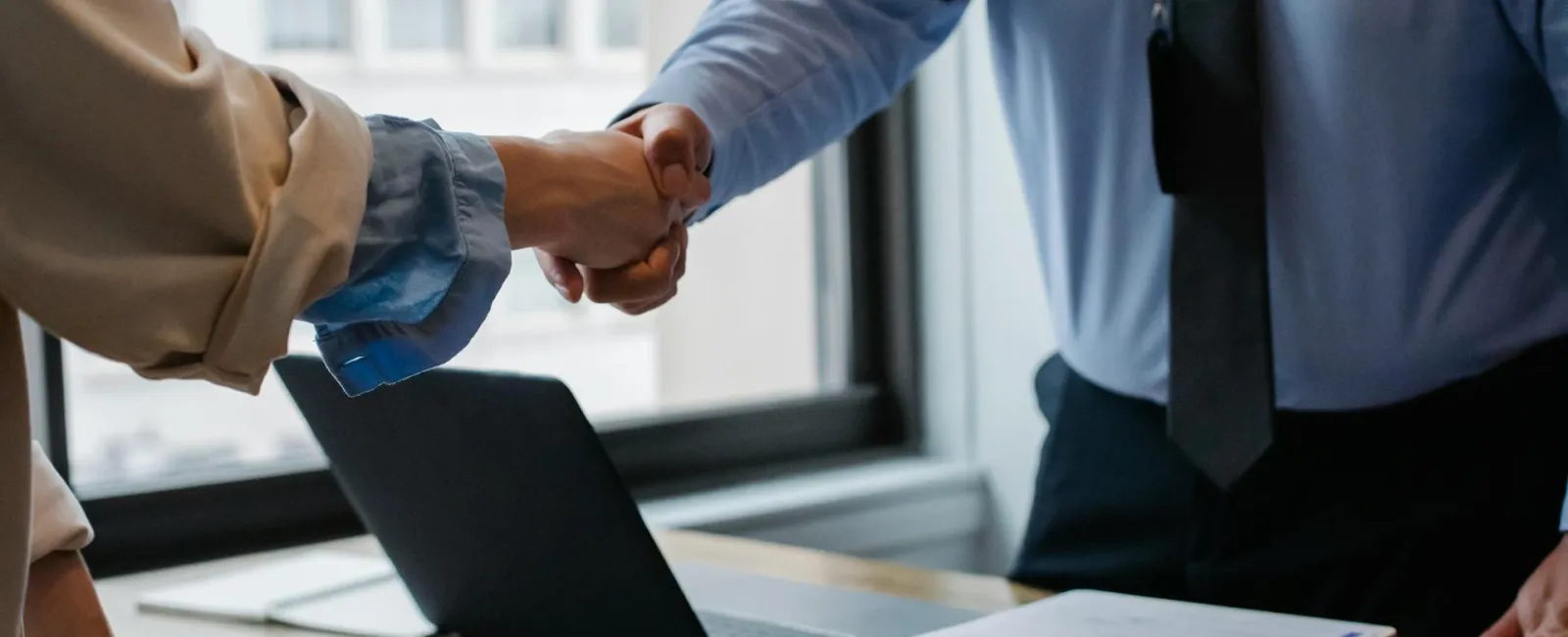 Two professionals shaking hands over a desk with a laptop and documents in a bright office setting