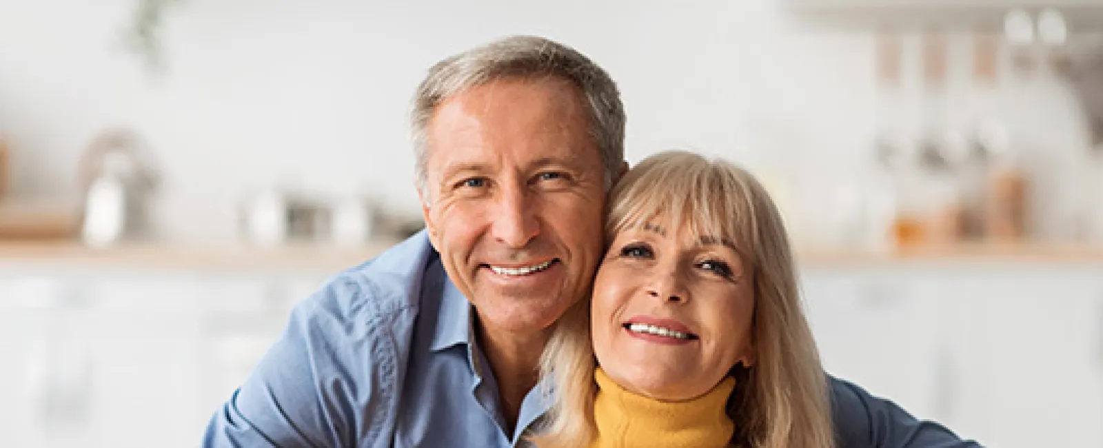 Smiling mature couple hugging on a couch in a cozy, modern living room with soft natural light.