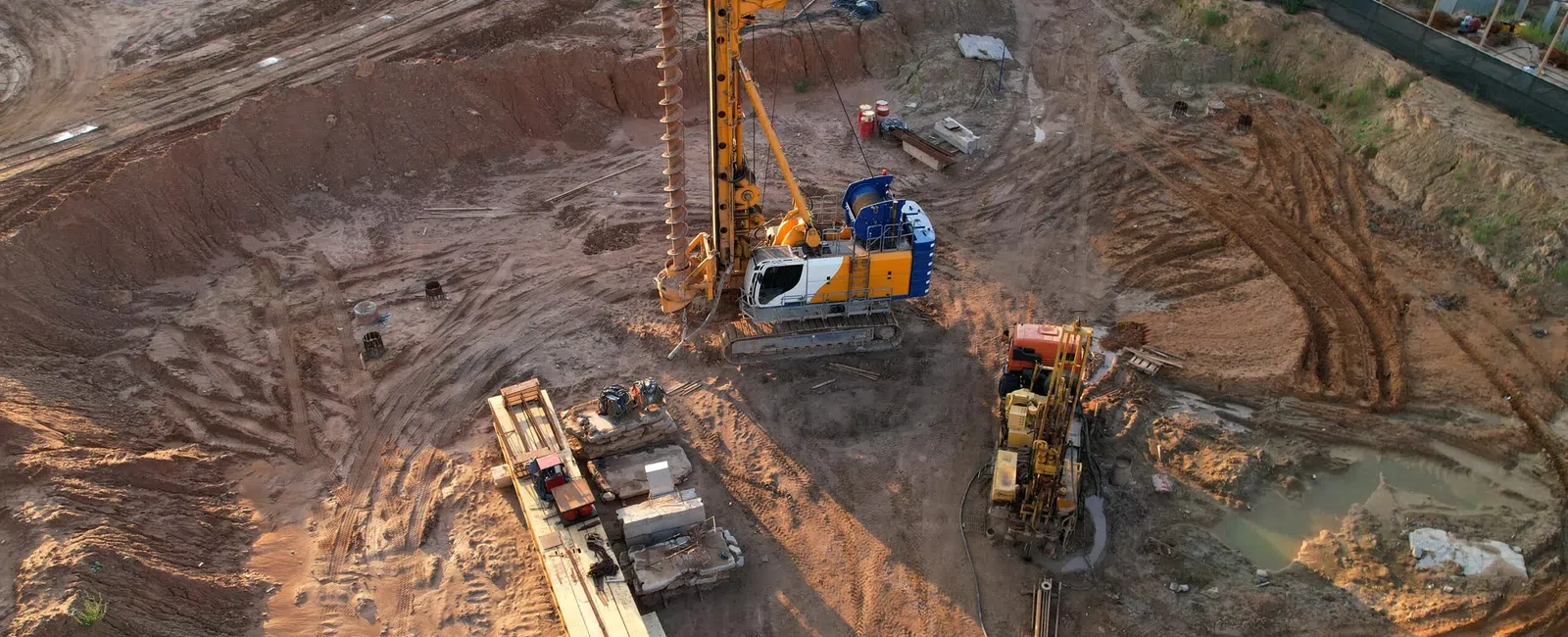 Aerial view of a construction site with heavy machinery drilling into the ground and building foundations nearby.