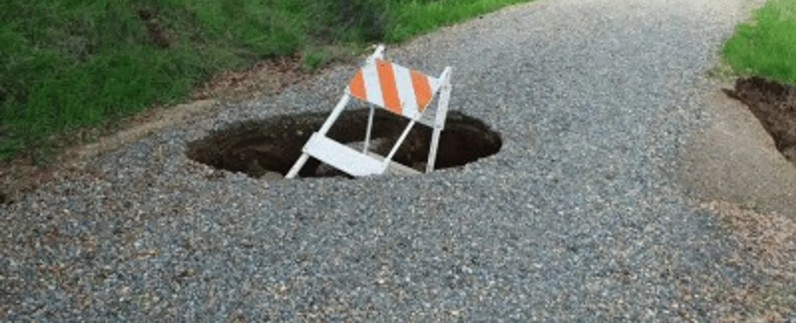 Gravel path with a large sinkhole containing a tilted orange and white barricade, surrounded by green grass.