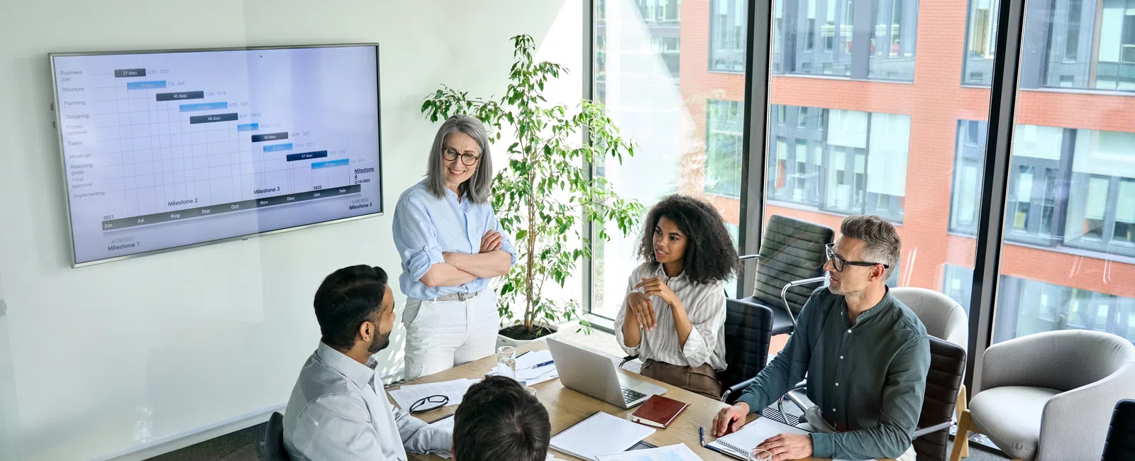 Team of diverse professionals discussing project timeline in modern office with large windows and digital screen.