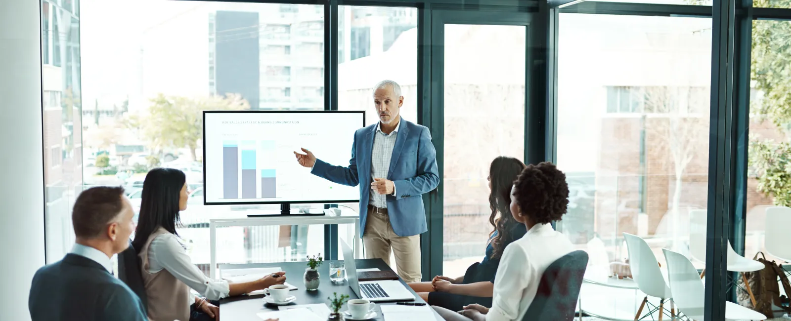 Business team in a modern conference room listening to a male presenter showing bar charts on a digital screen.