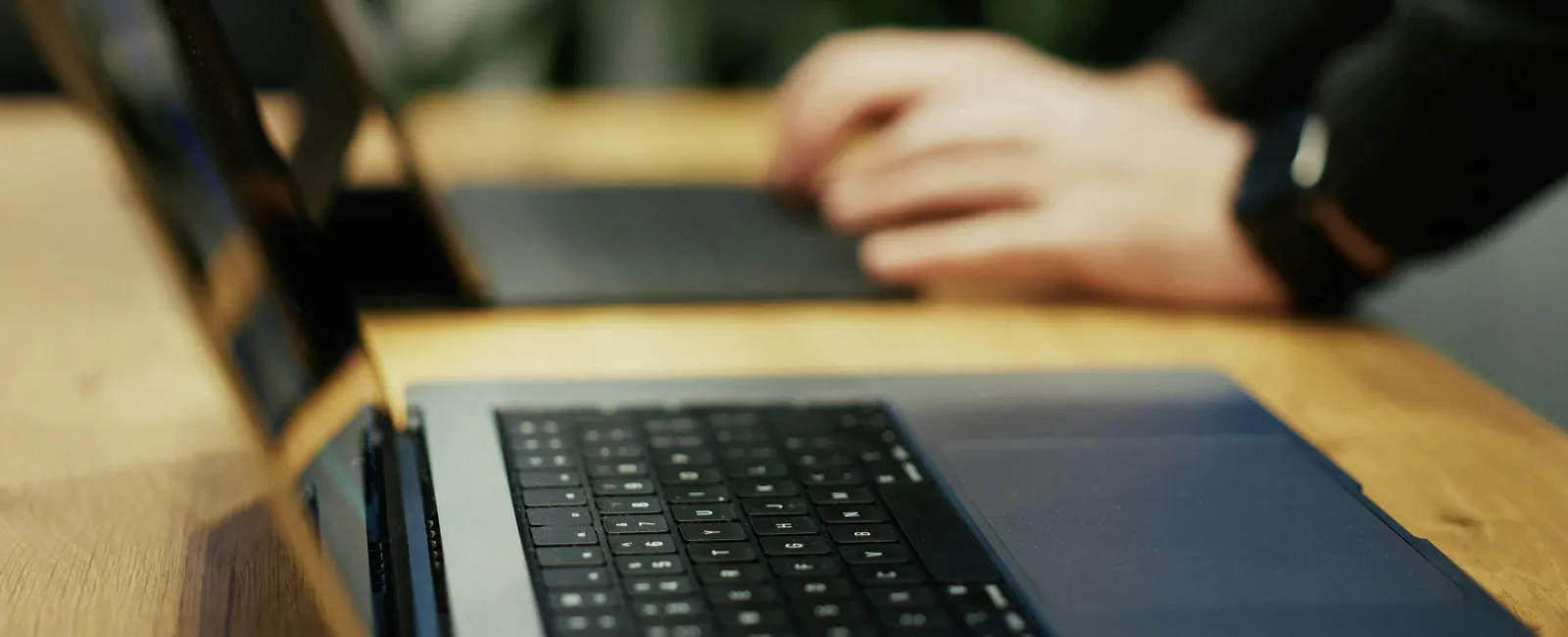 Close-up of a laptop keyboard on a wooden table with blurred hands typing in the background.