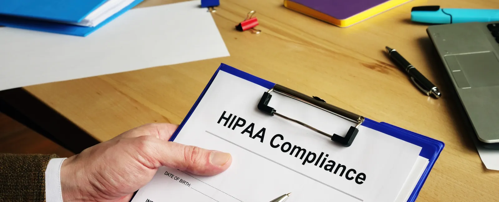 Person holding a HIPAA Compliance form on a clipboard with office supplies on a wooden desk.