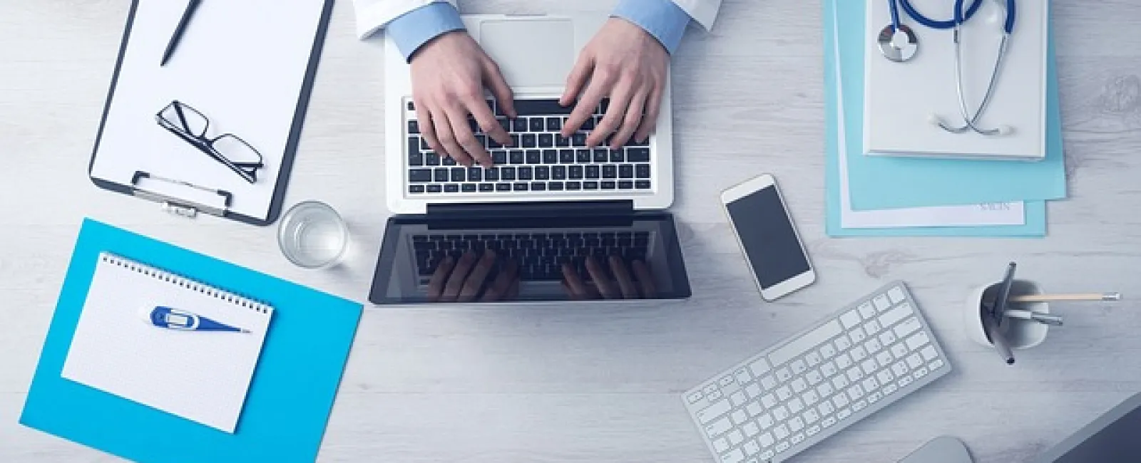 Doctor typing on a laptop surrounded by medical documents, a stethoscope, smartphone, and keyboard on a white desk.