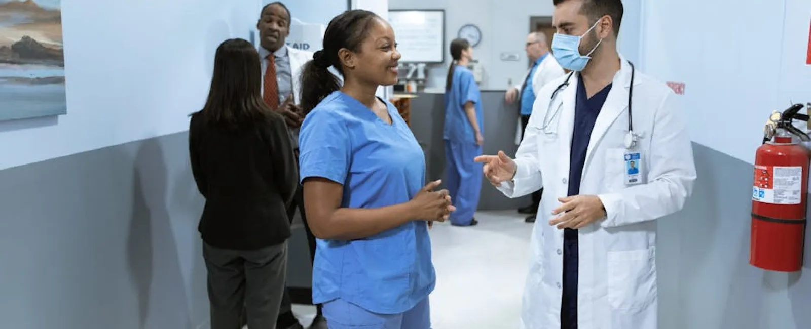 Doctor and nurse in conversation in hospital hallway with staff and medical equipment in background