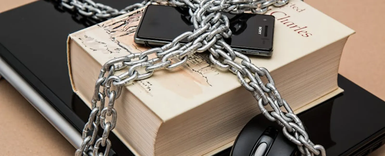 Thick chains wrapped around book, smartphone, mouse, and laptop symbolizing digital and information security.