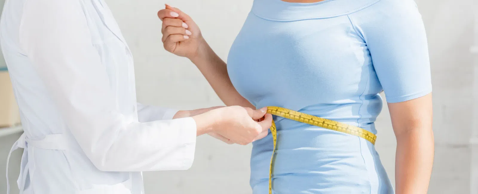 Healthcare professional measuring waist of woman in light blue shirt with yellow tape measure indoors.