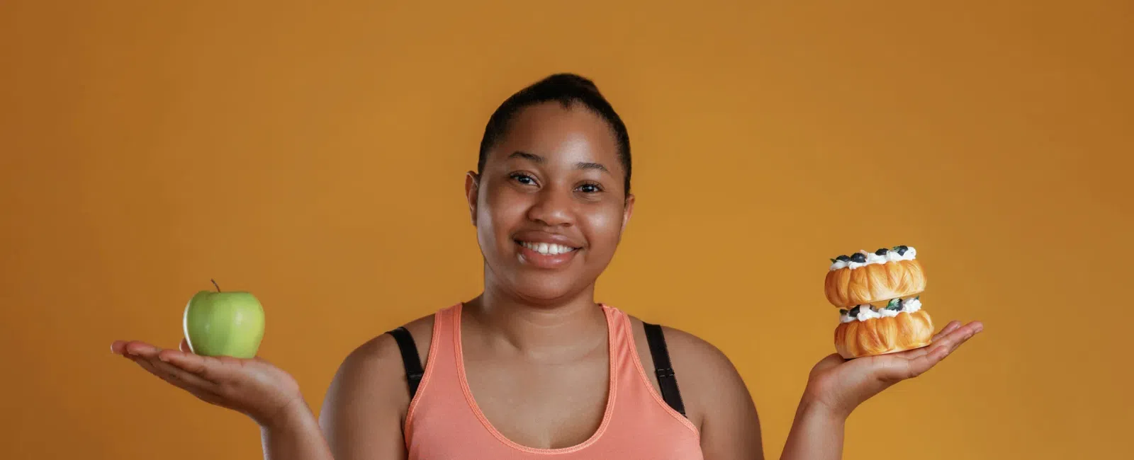 Smiling woman in peach tank top holds a green apple in one hand and a stack of mini desserts in the other against orange backdrop
