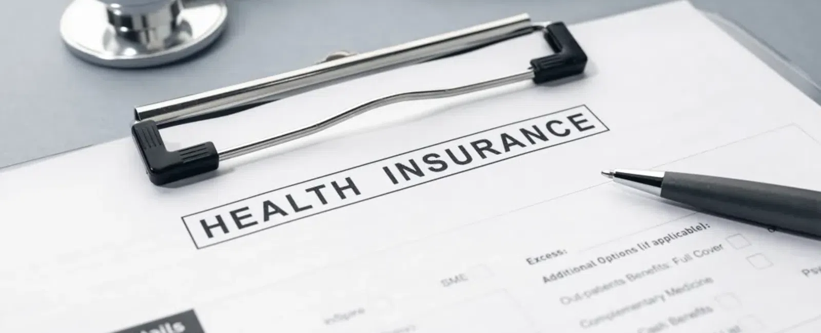 Health insurance form on clipboard with pen and stethoscope on a gray surface.
