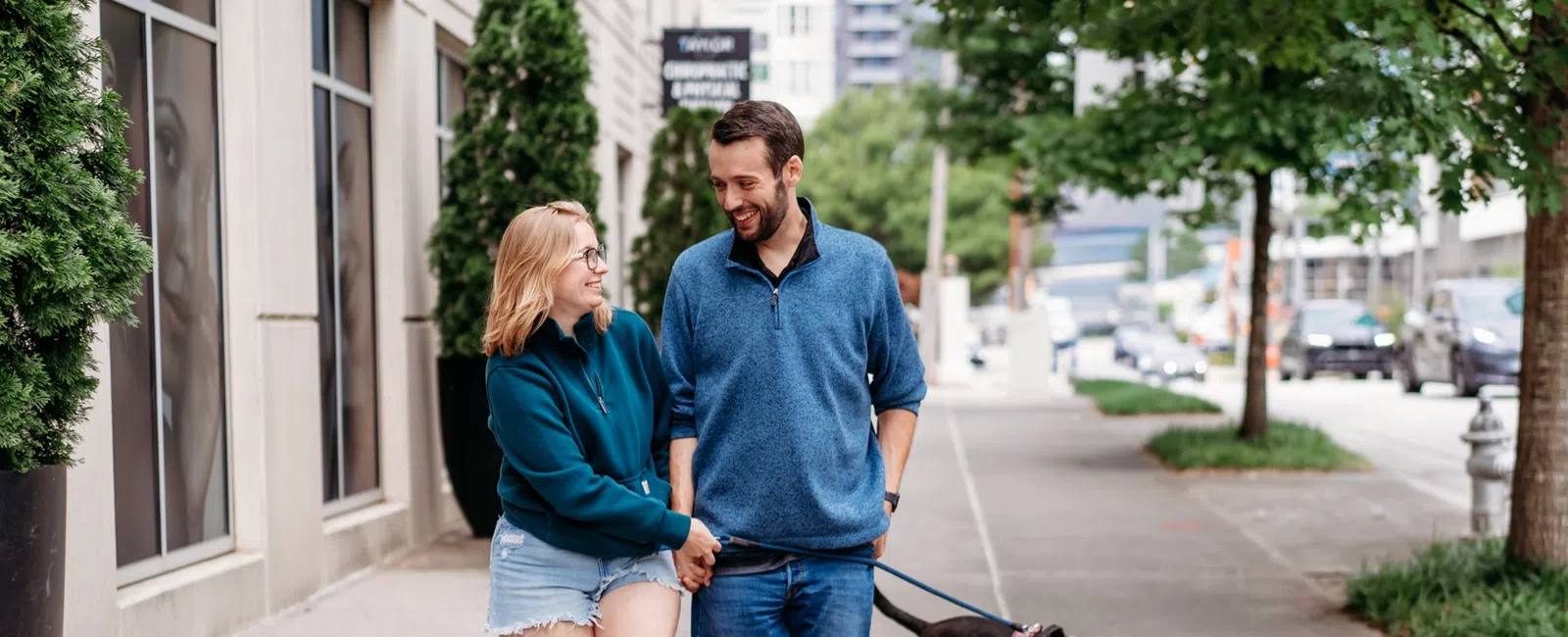 A couple walks their dog hand in hand along a tree-lined city sidewalk, illustrating the everyday outings people plan around after weight loss surgery.