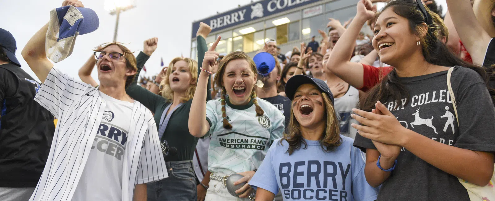 Berry College students cheer loudly at an outdoor event under evening stadium lights.