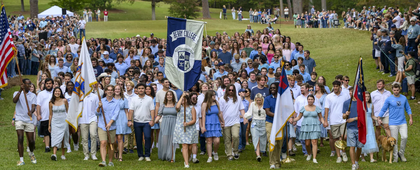 Large group of Berry College students walking outdoors holding flags during an organized event on green lawn.