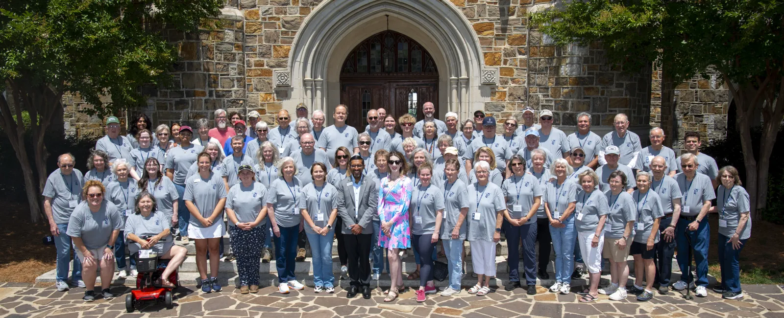 Large group of people wearing gray shirts posing in front of a stone building with an arched entrance under clear sunny sky