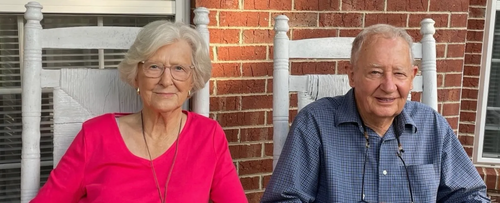 Elderly couple holding hands seated on white rocking chairs on a porch with a brick wall background.