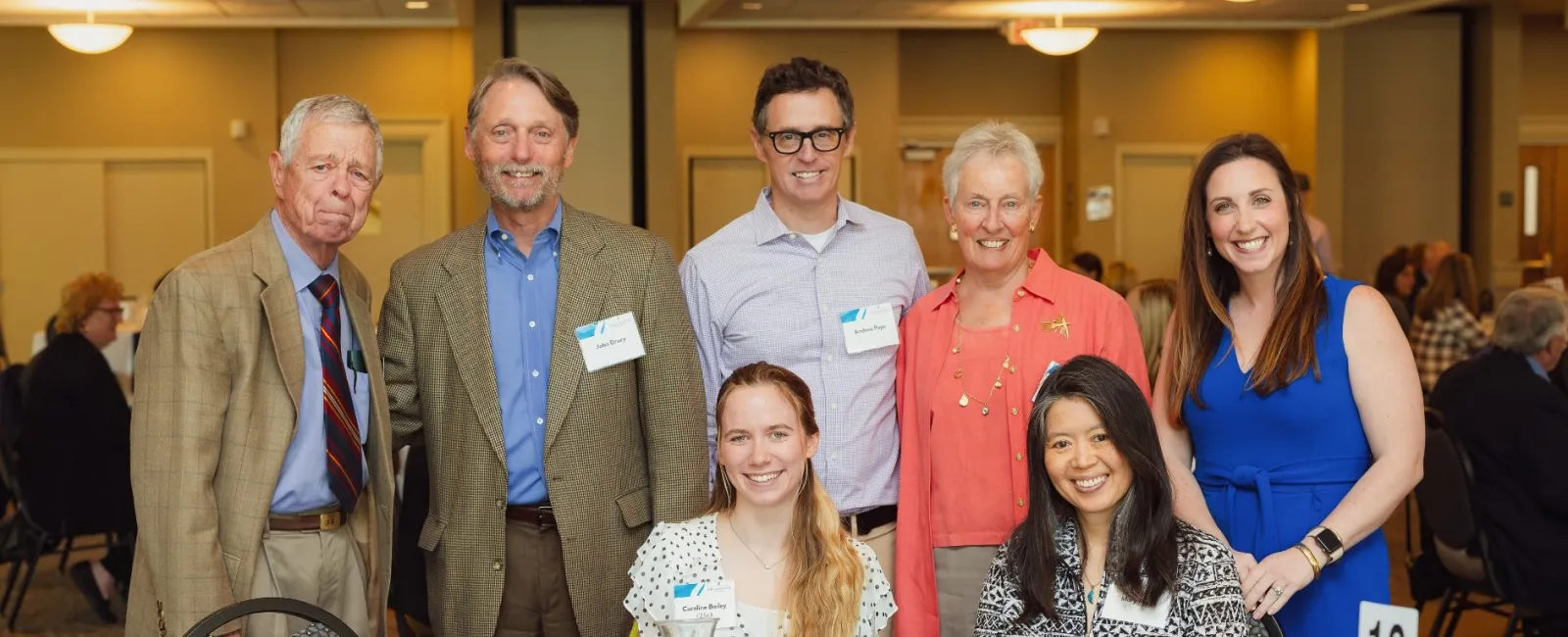 Seven diverse adults smiling at a banquet table with desserts and name cards in a conference room setting.
