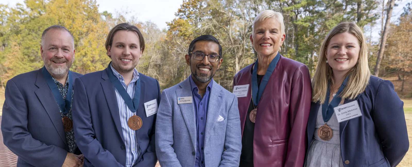 Five people wearing business attire and medals stand outdoors on a brick path with trees in the background.