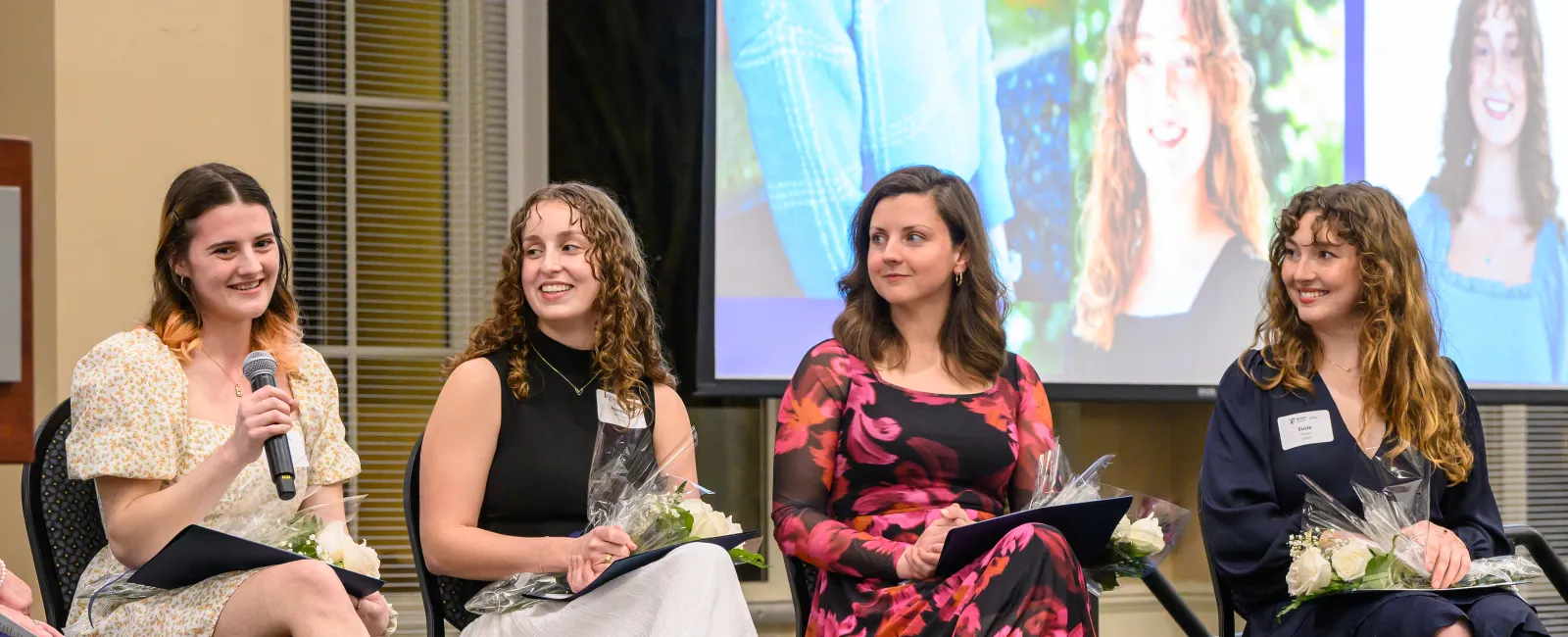Four women seated indoors at an event holding flowers, one speaking into a microphone with a projected image behind them
