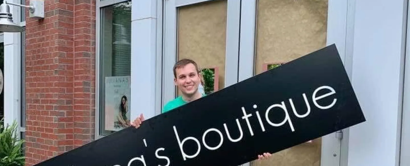 Man holding a large black sign with white text Juliana's boutique in front of a storefront with covered doors.