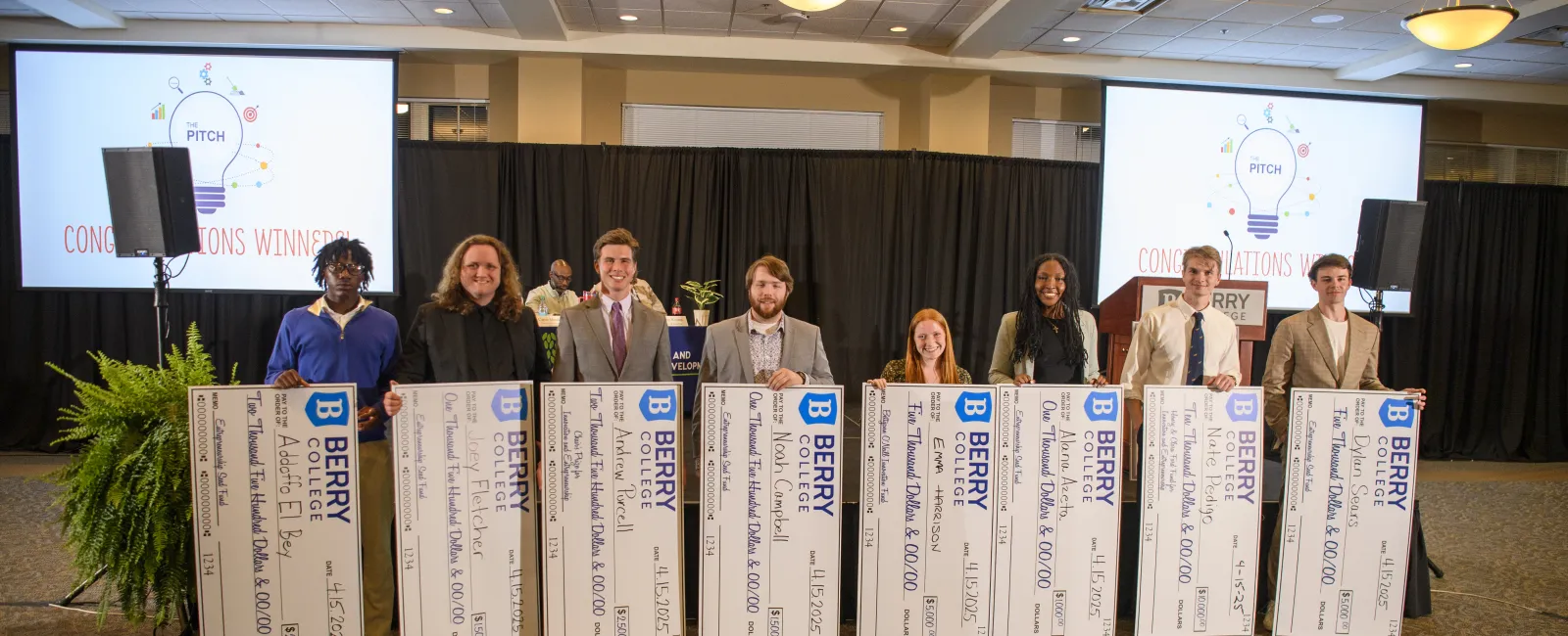 Group of eight diverse people holding oversized Berry College scholarship checks in a conference room.