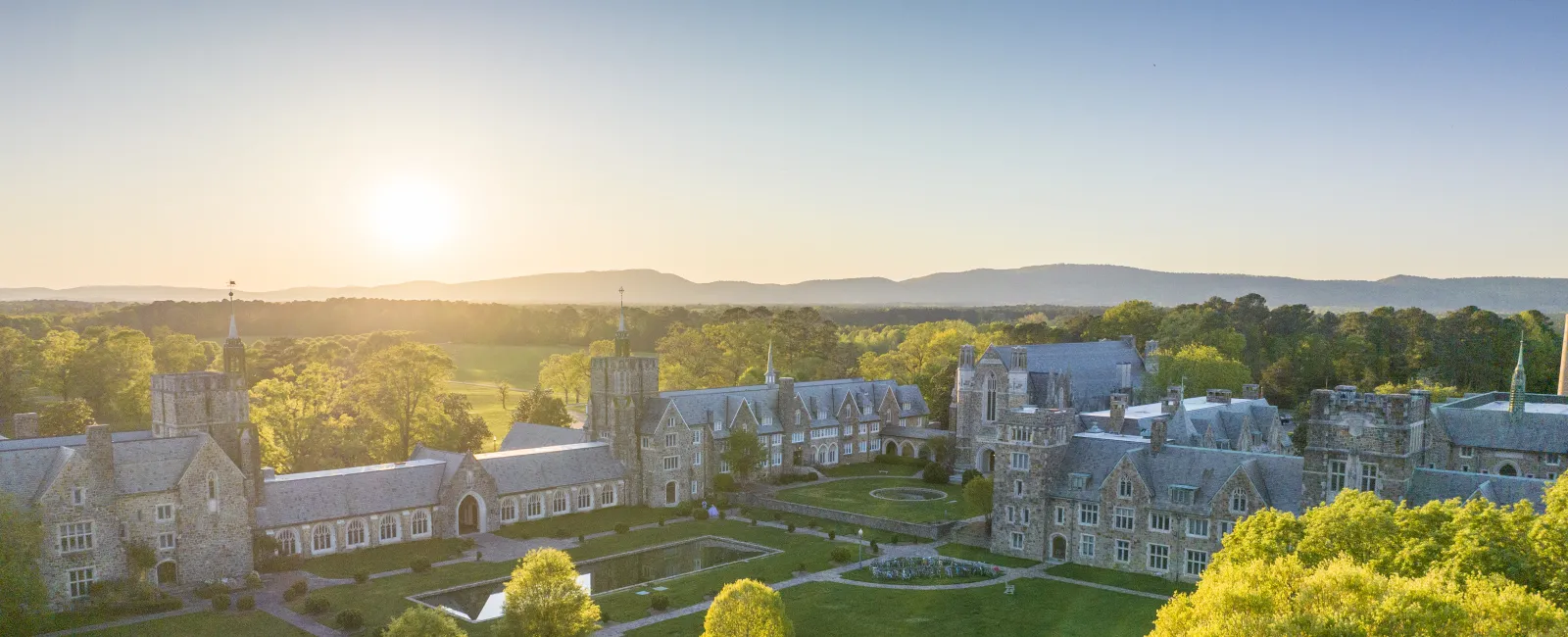 Aerial view of a historic college campus at sunrise, surrounded by lush greenery and scenic mountains.