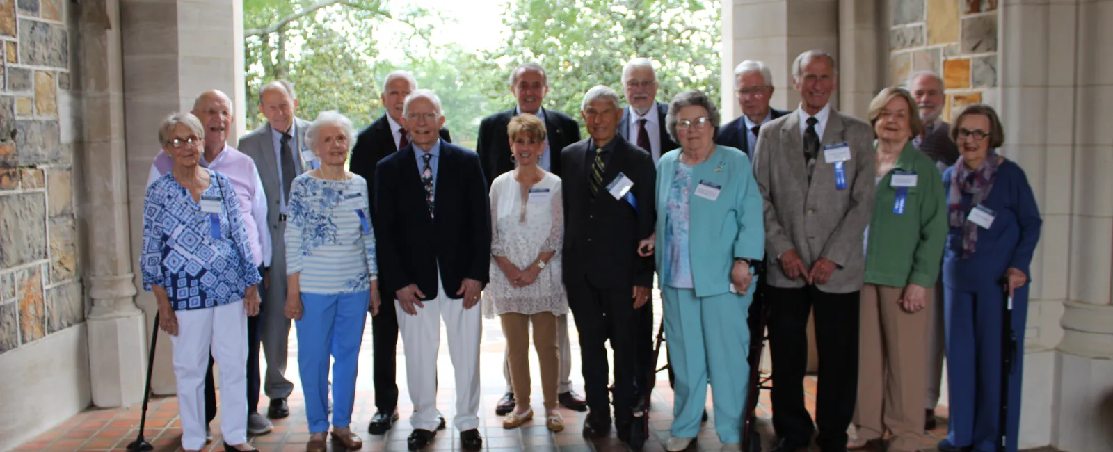 Group of elderly men and women standing in an archway with name tags, smiling at the camera during a reunion event.