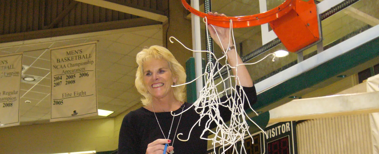 Woman on ladder cutting down basketball net after a game in a gymnasium with banners and scoreboard visible