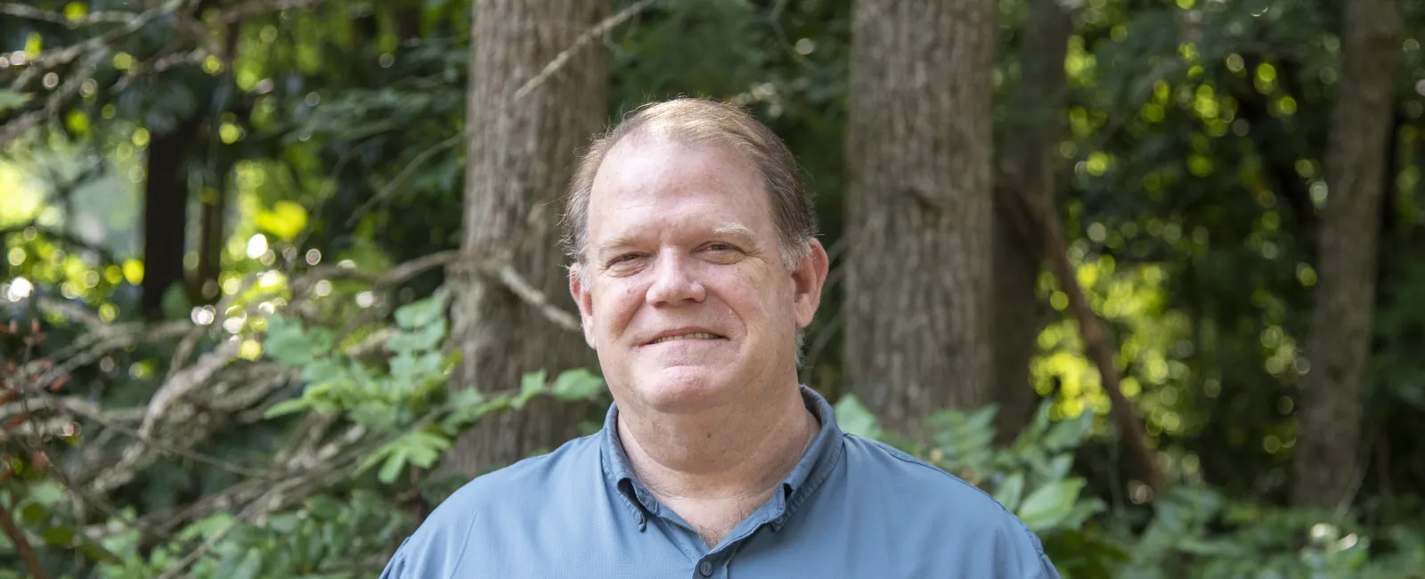 Middle-aged man smiling outdoors wearing a blue short-sleeve button-up shirt with trees and greenery in background