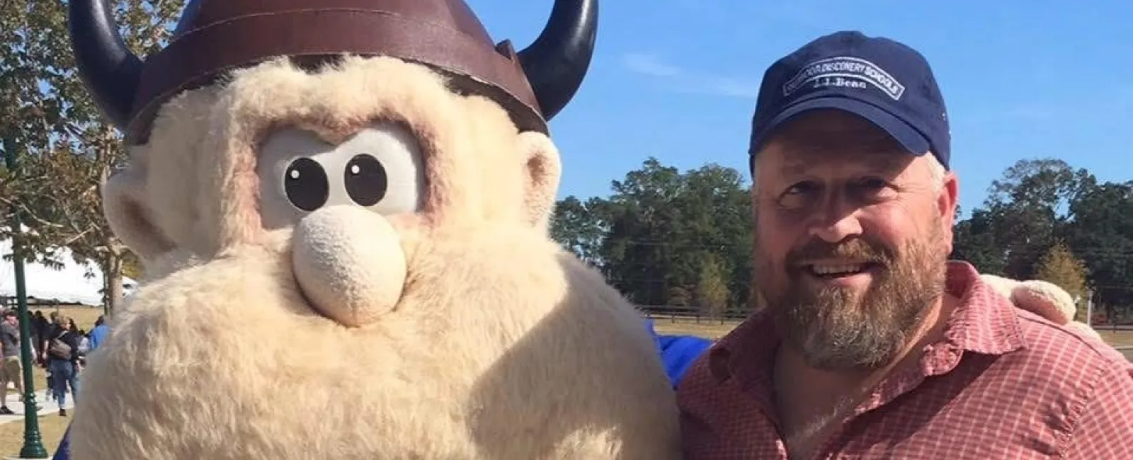 Man with beard posing outdoors next to large Viking mascot wearing horned helmet under blue sky