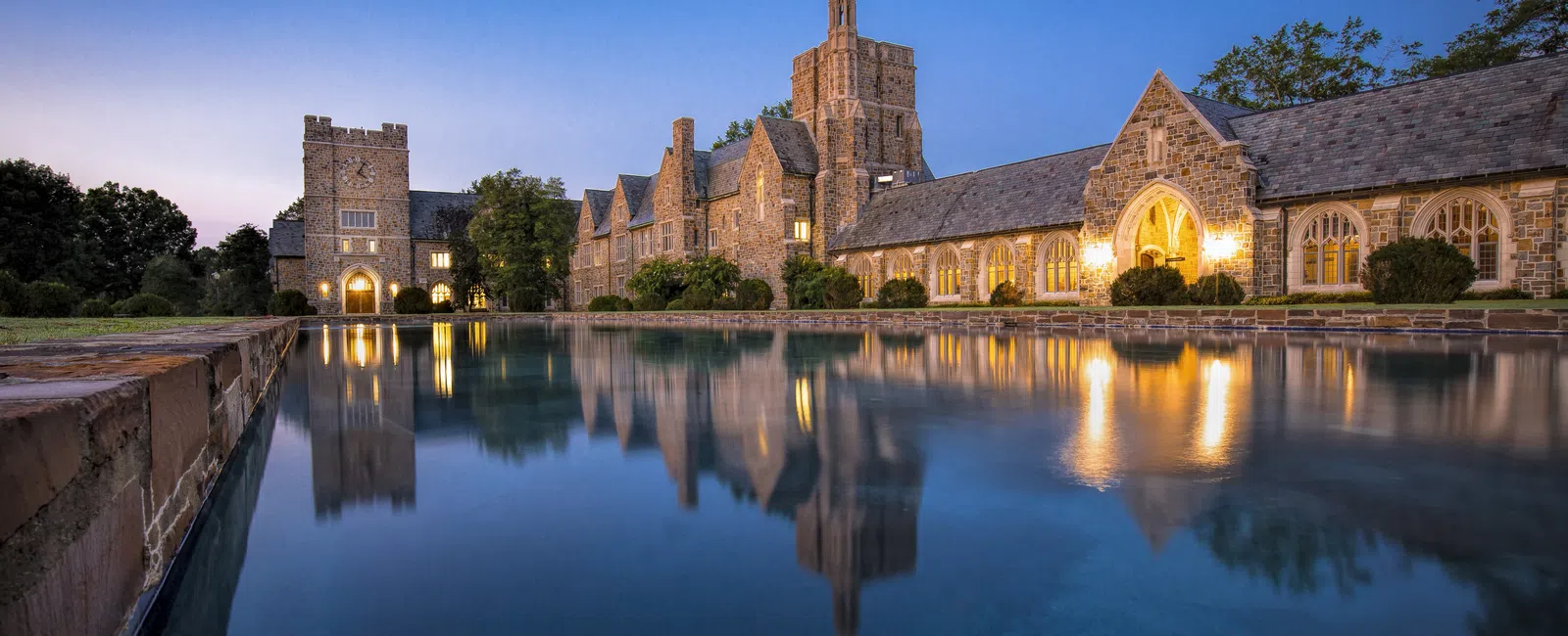 Historic stone building reflected in a calm water pool under a clear twilight sky with warm lights.