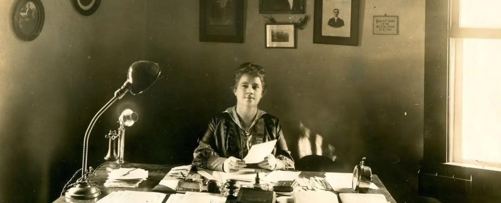 Early 20th-century woman sitting at desk with papers and vintage telephone under portraits on wall