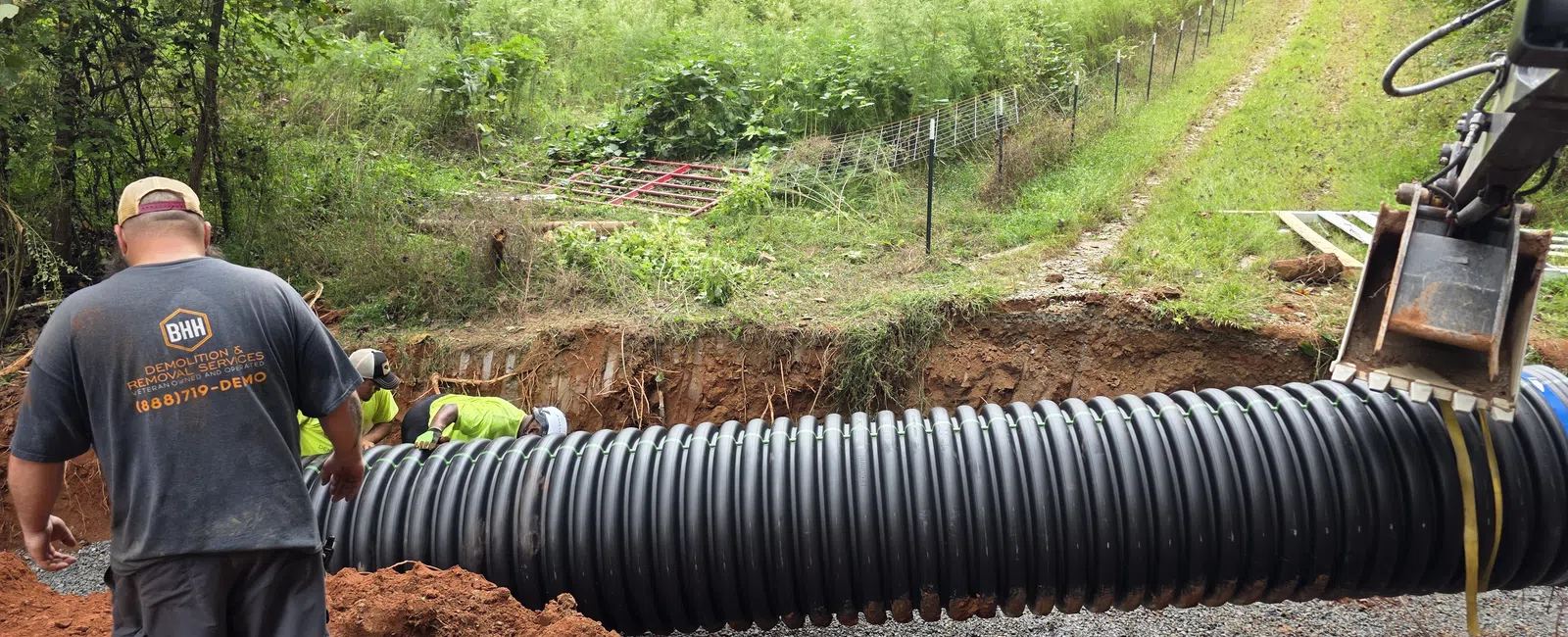Workers installing large black corrugated drainage pipe in a rural area near a mobile home and dirt road.