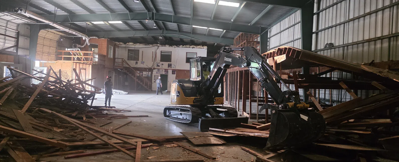 Indoor construction site with excavator clearing wooden debris inside a large warehouse with workers present