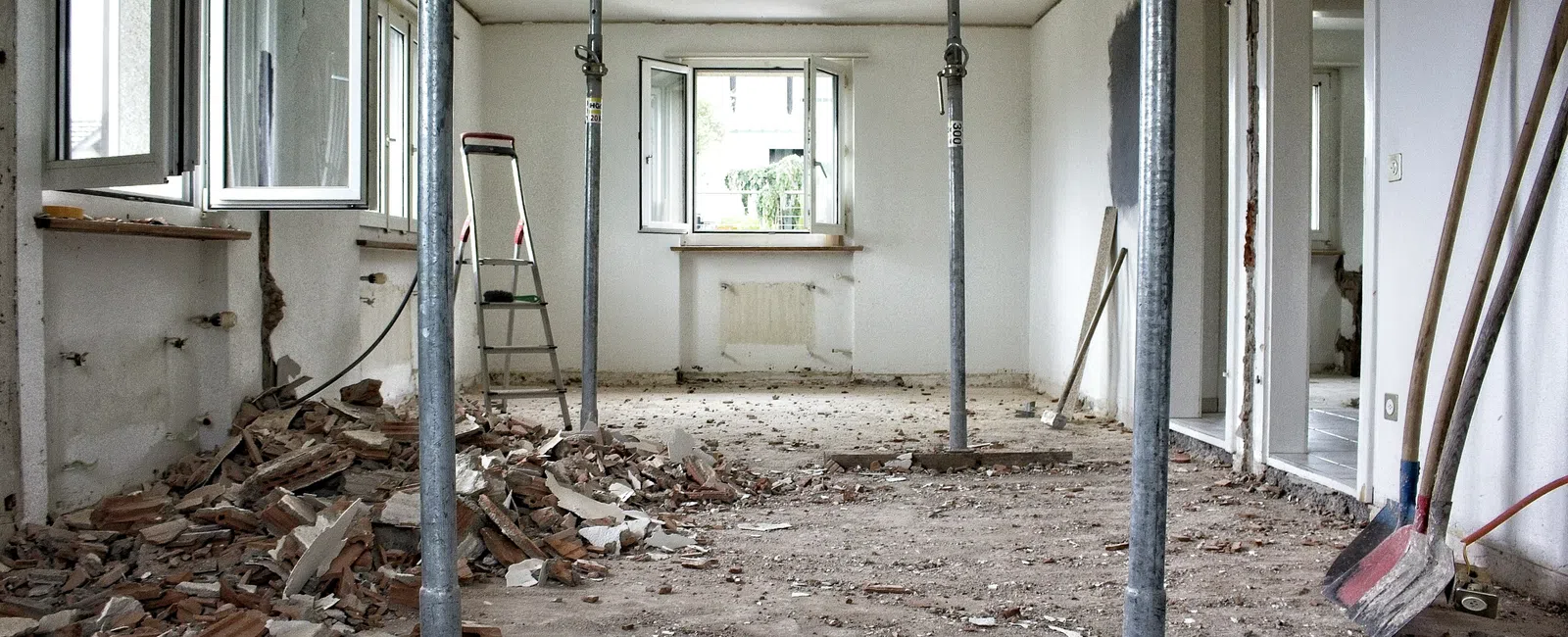 Room under renovation with open windows, scattered debris, metal support pillars, and construction tools visible.