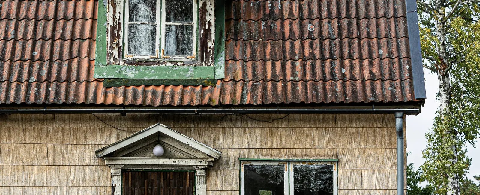 Old beige house with weathered red tile roof, peeling paint, a broken window, and a pink scooter outside.
