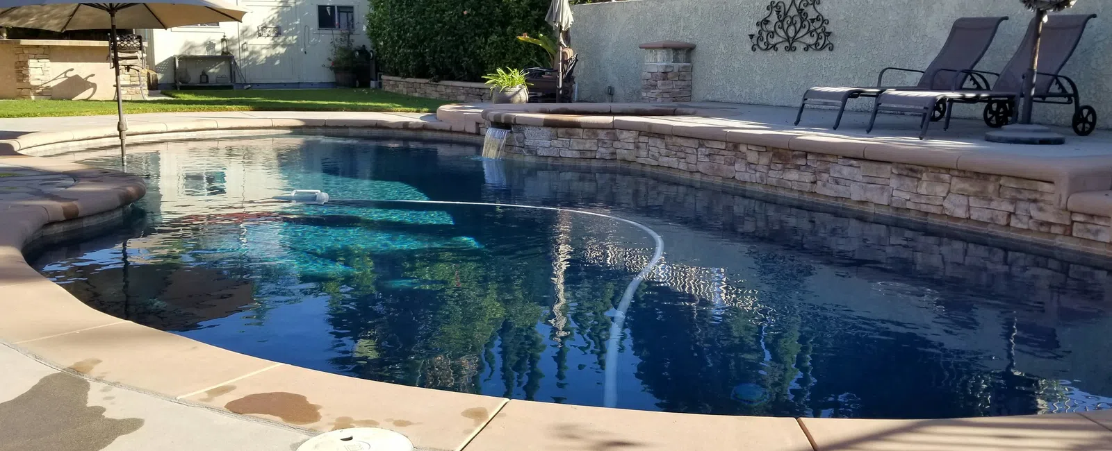 Clear blue backyard swimming pool with stone wall, lounge chairs, and large umbrella on sunny day.