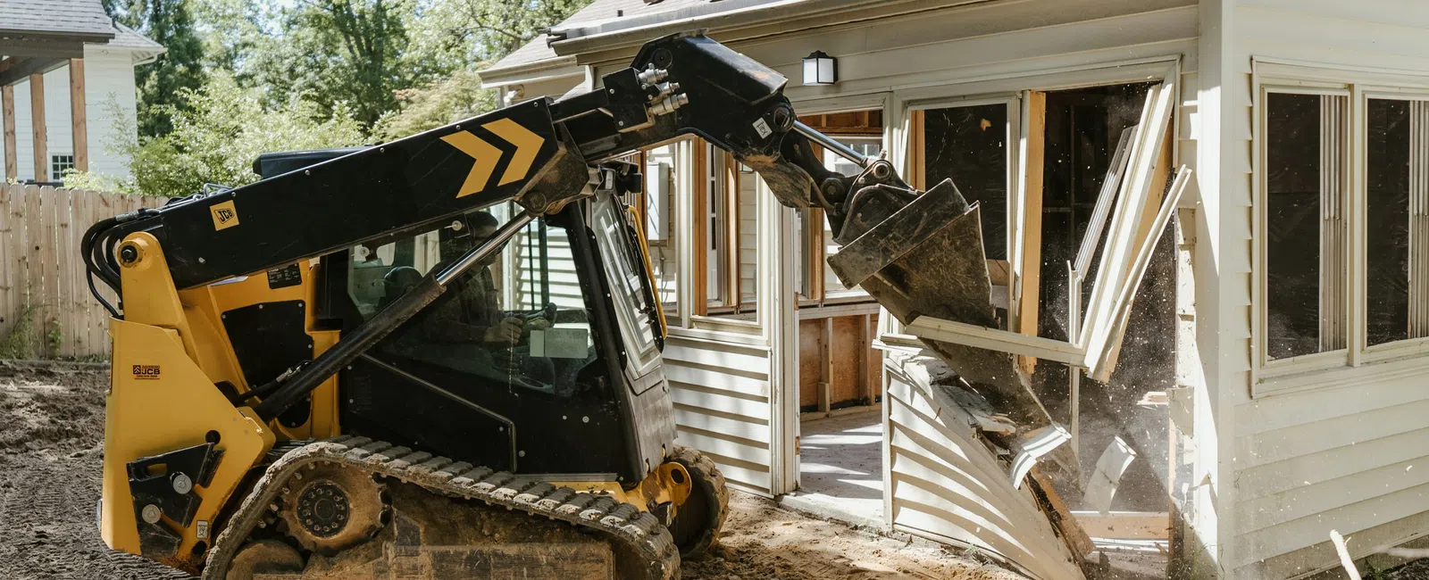 Yellow skid steer loader demolishing a house wall on a residential construction site with dirt and debris.