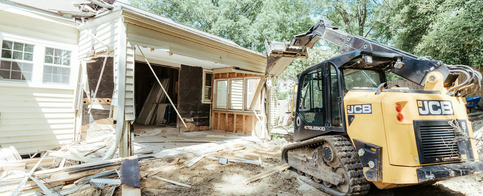 Yellow JCB excavator demolishing a house with debris and exposed interior under bright daylight.