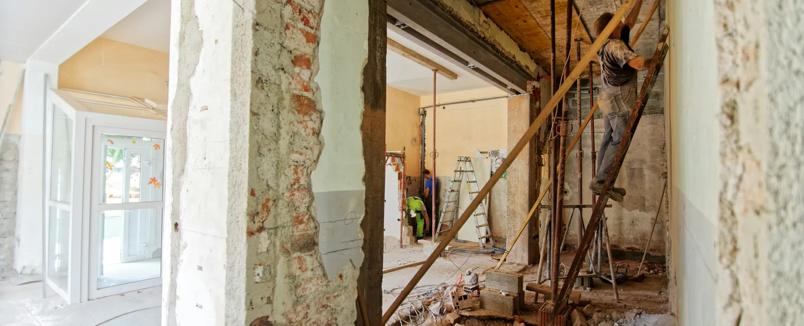 Construction worker repairs interior wall framework in a building renovation site with exposed bricks and debris.