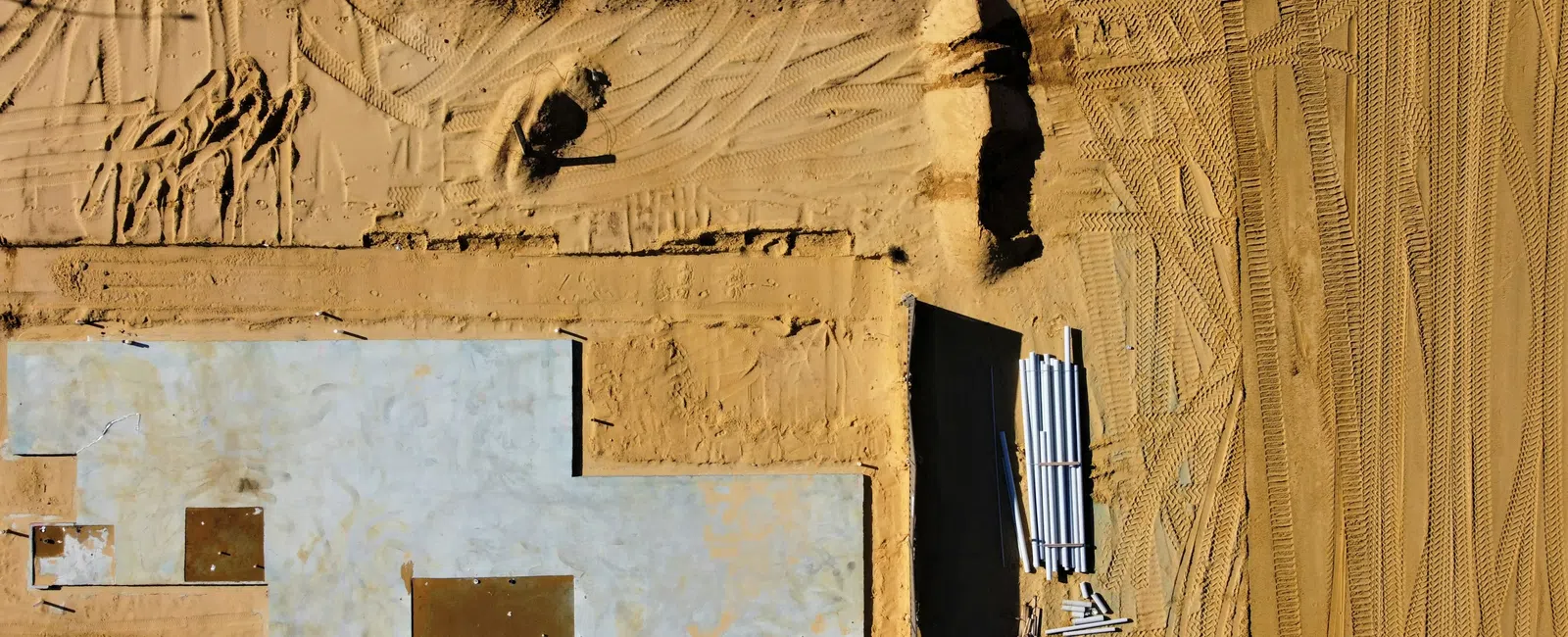 Aerial view of a construction site showing a concrete foundation on sandy ground with tire tracks.