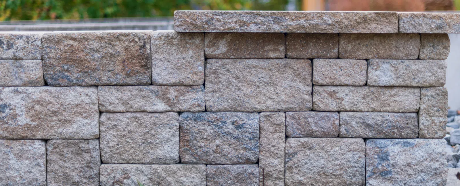 Close-up of a beige stacked stone retaining wall with garden soil and plants in the foreground and blurred greenery behind.