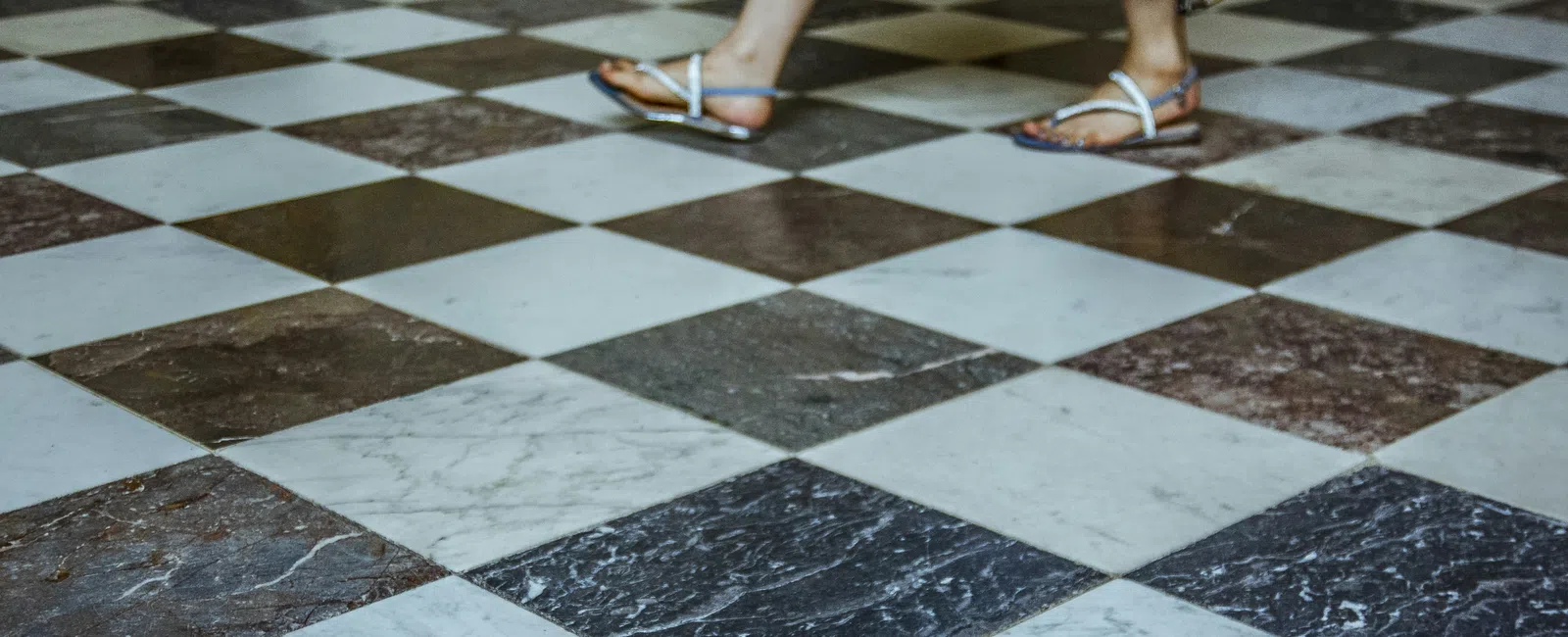 Person wearing white sandals walks on large black and white checkered marble floor with patterned dress.