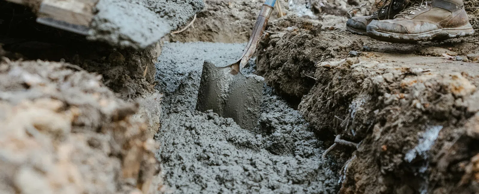 Worker pouring and leveling wet concrete into a trench with a shovel at a construction site outdoors.
