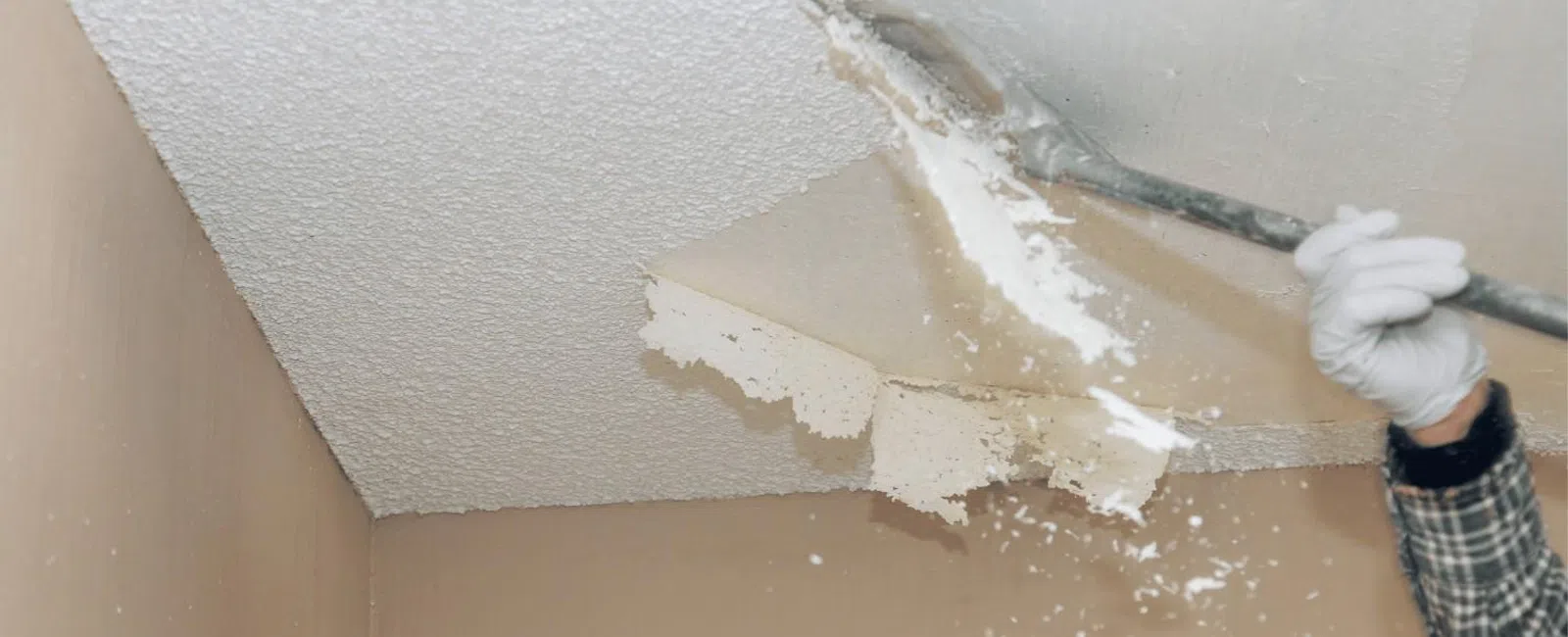Person removing popcorn ceiling texture with a scraping tool and gloved hand in a beige room.