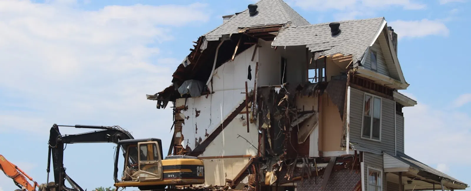 Partially demolished two-story house with construction machinery and debris under a blue sky.