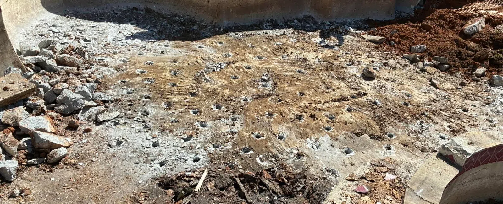 Partially demolished concrete swimming pool with exposed dirt and broken debris inside under sunlight.