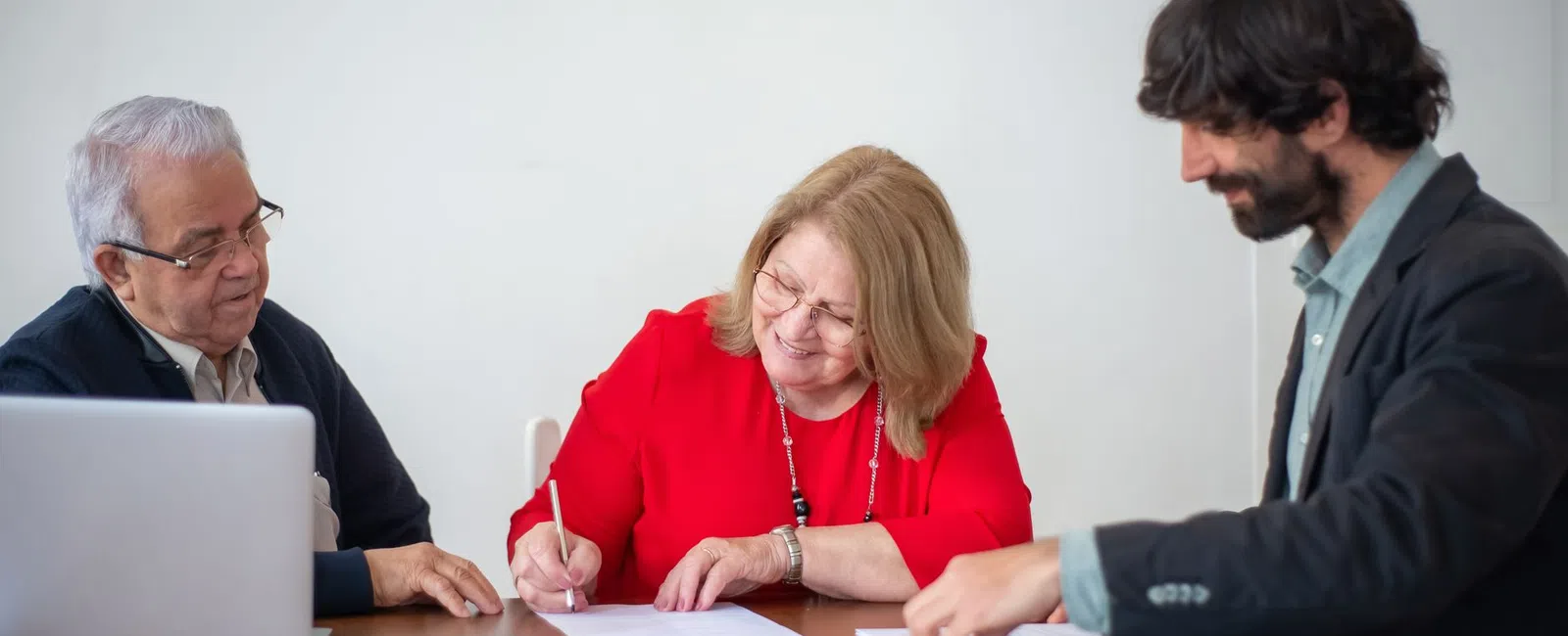 Three professionals at a table signing documents and discussing work with a laptop and tablet present