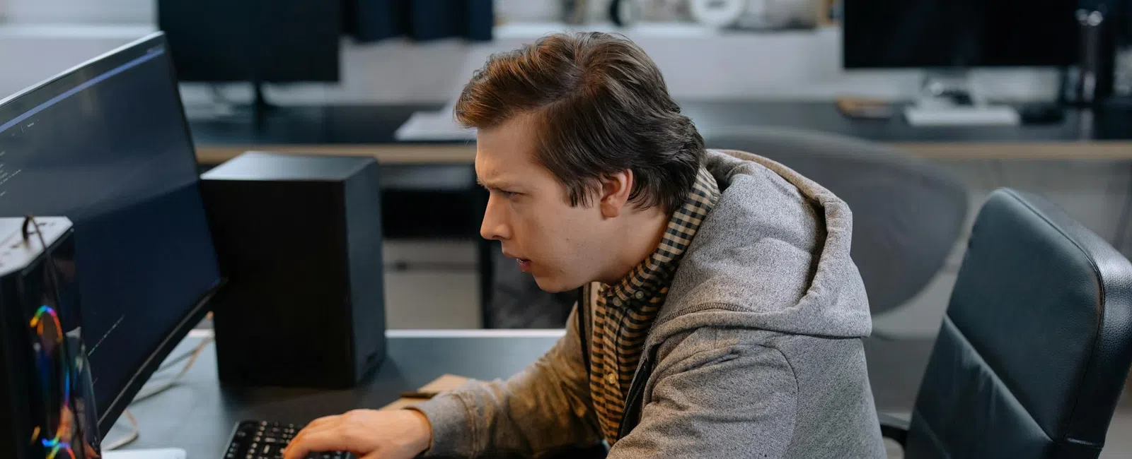 Young man wearing a gray hoodie working intensely on a desktop computer in an office setting.