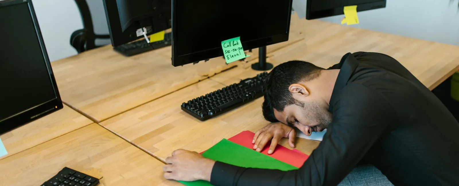 Man asleep at desk with computers, resting head on colorful folders in a modern office setting.