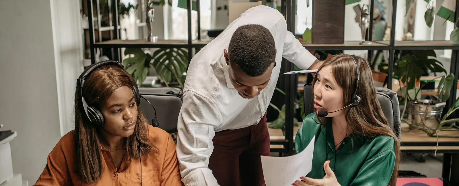Diverse team discussing work with laptops and headsets in a modern office filled with plants and documents.